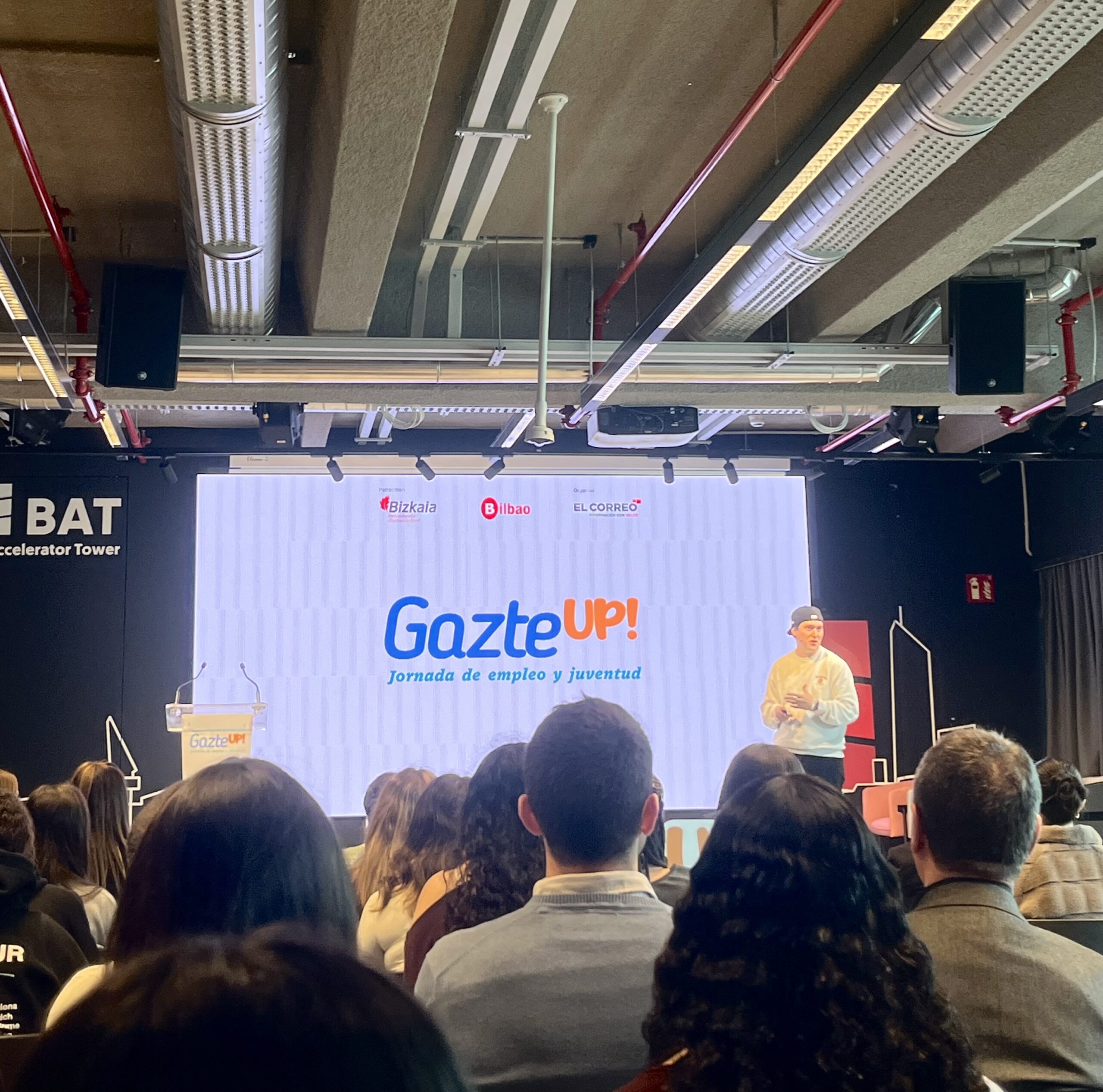 GazteUP! enplegu eta gazteria jardunaldiaren irudia BAT B Accelerator Tower-en. Hizlari bat agertoki gainean dago azalpenak ematen pantaila handi baten aurrean, eta publikoa bizkarrez ikusten da areto moderno eta industrial batean.